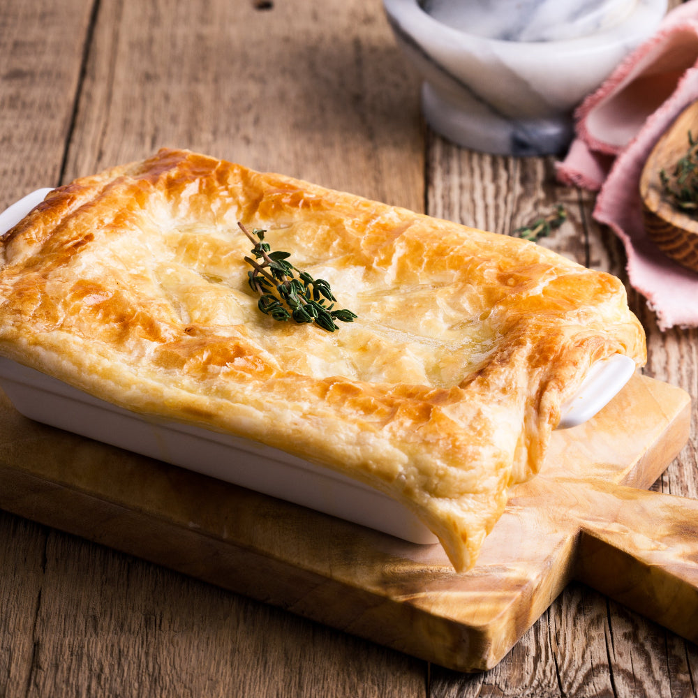 A baked rectangular pie or casserole with a golden flaky crust sits on a wooden cutting board on a rustic wooden table. A small sprig of fresh thyme is placed on top, with blurred dining props in the background.