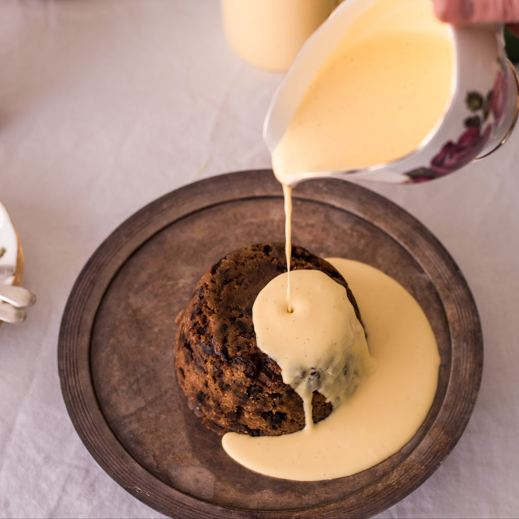 Custard being poured over a dessert on a wooden plate with a glass of juice and teacup in the background.